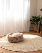 Solace Aura beige cat bed made from natural merino wool, styled on a round jute rug in a sunlit interior with sheer curtains, wooden floor, woven plant basket and chair in the background, photographed from a wide lifestyle angle