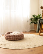 Solace Aura Cappuccino merino wool cat bed in warm brown tones, handmade round cat bed placed on a natural jute rug in a sunlit living room with sheer white curtains, timber floor, indoor plant and wooden chair in the background