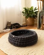 Solace Aura charcoal Merino wool cat bed made from natural wool, photographed in a warm minimalist home with timber floors, jute rug, and a relaxed cat in the background.