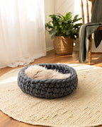 Solace Aura Moonstone grey merino wool cat bed resting on a round jute mat on timber floorboards, with a relaxed cat curled inside the bed. Soft white curtains filter natural light beside a leafy indoor plant in a woven pot and a wooden chair draped with a knitted throw, creating a calm, earthy interior setting.