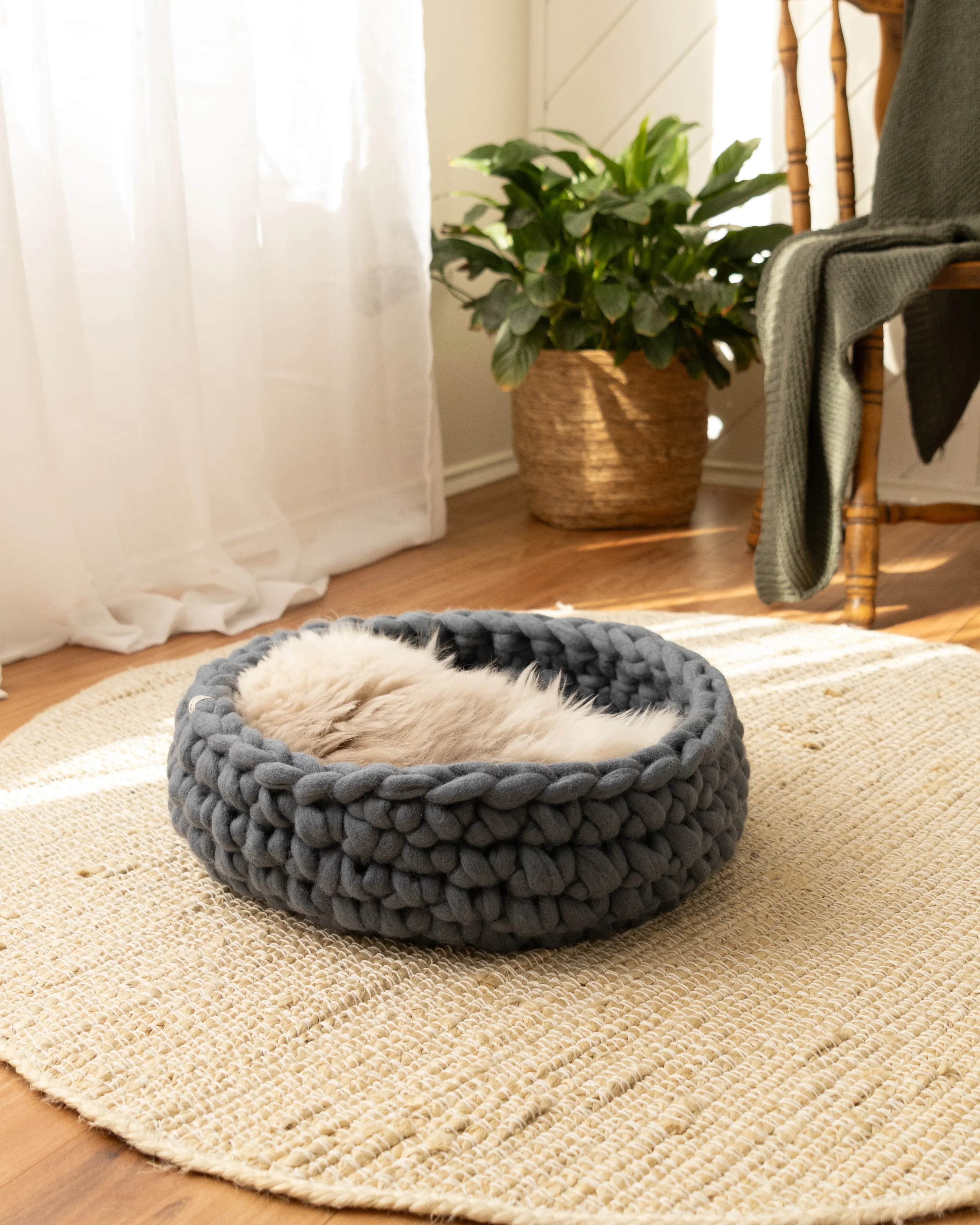 Solace Aura Moonstone grey merino wool cat bed resting on a round jute mat on timber floorboards, with a relaxed cat curled inside the bed. Soft white curtains filter natural light beside a leafy indoor plant in a woven pot and a wooden chair draped with a knitted throw, creating a calm, earthy interior setting.