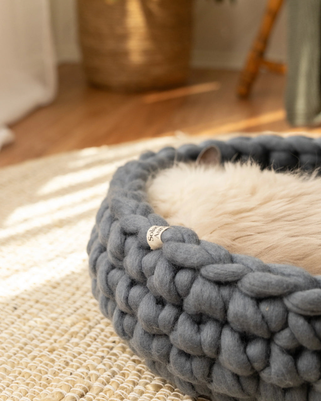 Solace Aura Moonstone grey merino wool cat bed styled on a natural jute rug over timber floors, with soft white curtains, a leafy plant in a woven basket, and a wooden chair draped with a knit throw in the background.