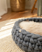 Solace Aura Moonstone grey merino wool cat bed styled on a natural jute rug over timber floors, with soft white curtains, a leafy plant in a woven basket, and a wooden chair draped with a knit throw in the background.