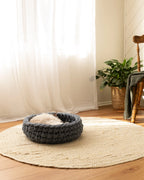 Solace Aura Moonstone grey merino wool cat bed placed on a natural jute rug over timber floorboards, with a cat resting inside. Soft white curtains, a leafy indoor plant in a woven basket, and a wooden chair with a draped blanket create a calm, earthy interior setting.