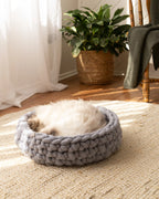 Solace Aura Pebble Grey cat bed made from natural merino wool, photographed on a woven jute rug in a softly lit home interior, with a fluffy cat resting comfortably inside the bed, sheer curtains, timber floor, and a leafy indoor plant in the background.