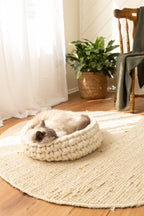 Solace Aura Natural White cat bed made from merino wool, featuring a cat resting inside the bed on a round jute mat in a sunlit living space with sheer curtains, wooden floor, indoor plant, and timber chair in the background.