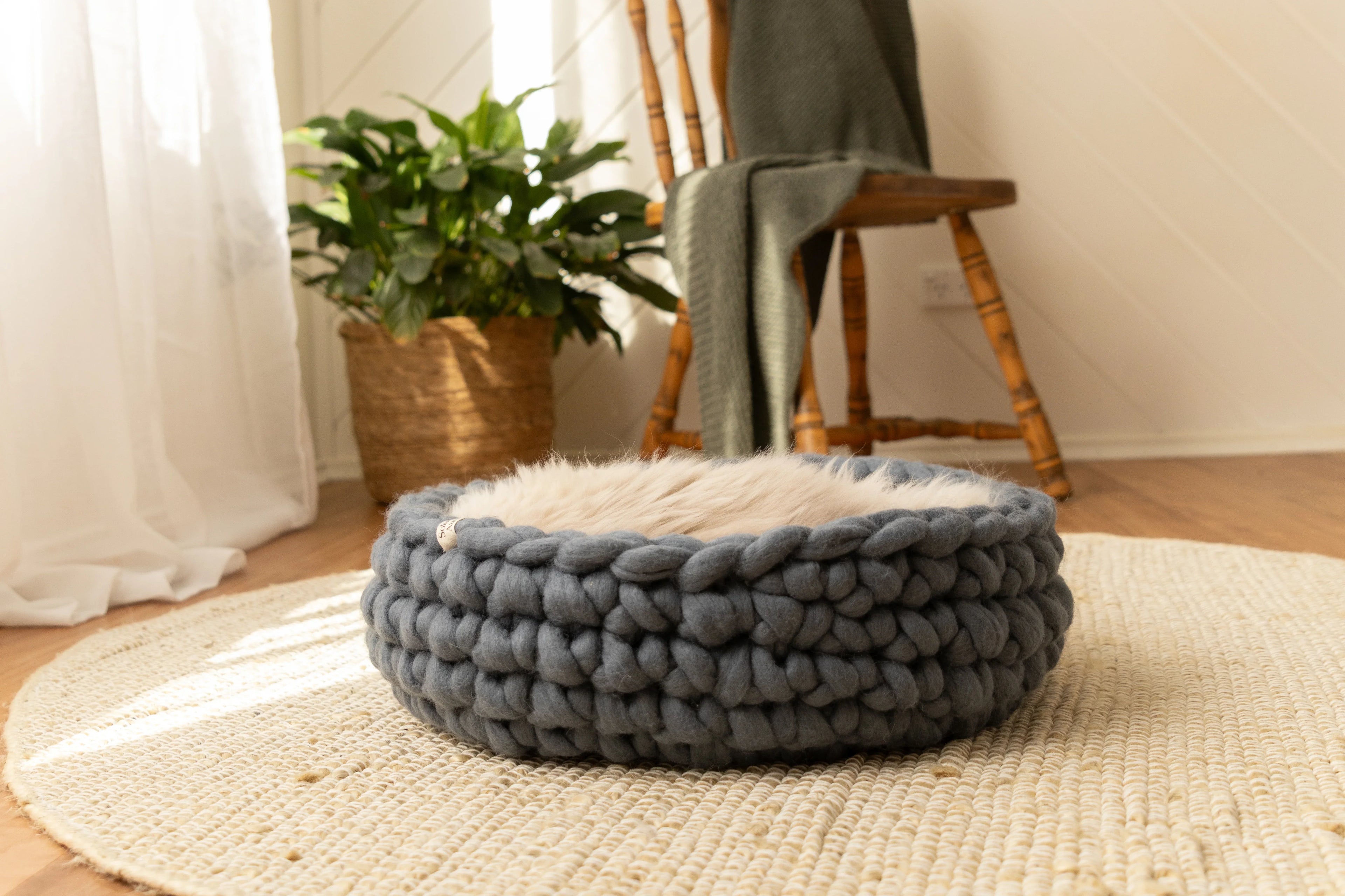 Solace Aura Moonstone grey merino wool cat bed styled on a natural jute rug over timber floors, with soft white curtains, a leafy plant in a woven basket, and a wooden chair draped with a knit throw in the background.