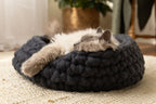 Charcoal grey Solace Aura cat bed handcrafted from 100% merino wool, with a cat resting inside on a neutral woven rug in a sunlit home.