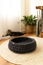 Solace Aura charcoal Merino wool cat bed made from natural wool, photographed in a warm minimalist home with timber floors, jute rug, and a relaxed cat in the background.