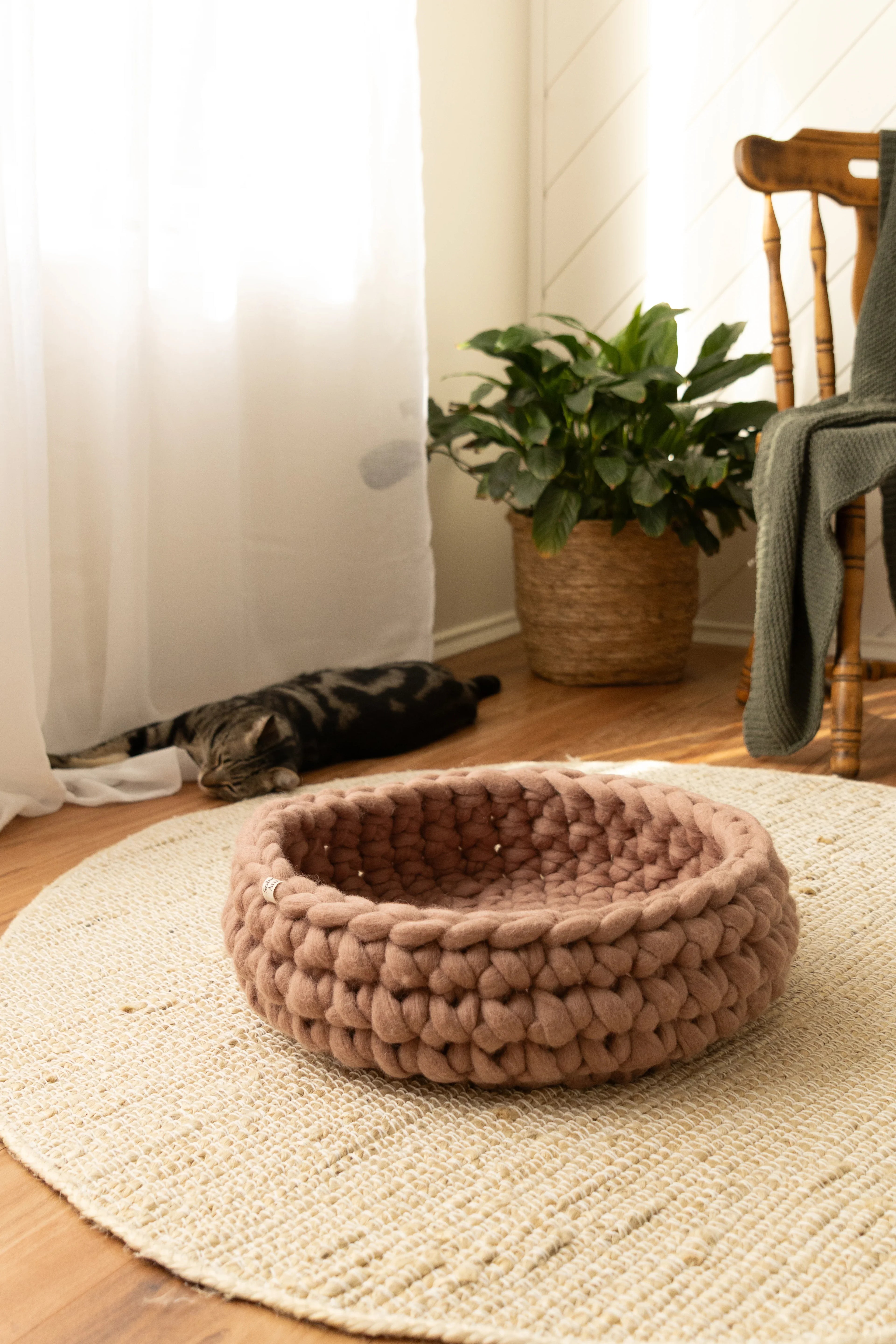 Solace Aura Cappuccino cat bed in warm brown tone, handmade merino wool cat bed, styled on a natural jute rug with soft daylight, featuring a woven round bed in the foreground and a relaxed cat resting near sheer curtains in a calm, earthy home interior.