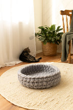Solace Aura Pebble Grey merino wool cat bed styled on a round jute rug over timber floorboards, with soft white curtains, a woven plant pot, tabby cat beside the bed and a wooden chair creating a calm, natural interior setting.