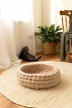 Solace Aura Beige cat bed made from natural merino wool, styled on a round jute rug in a softly lit home interior, with sheer curtains, timber flooring, a potted plant, tabby cat and a wooden chair in the background