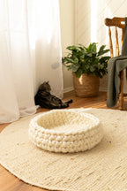 Solace Aura Natural White cat bed made from merino wool, styled on a round jute rug in a sunlit interior, with the empty cat bed in the foreground and a relaxed tabby cat resting near sheer curtains, a woven plant basket, and a wooden chair in the background