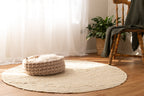 Solace Aura beige cat bed made from natural merino wool, styled on a round jute rug in a sunlit interior with sheer curtains, wooden floor, woven plant basket and chair in the background, photographed from a wide lifestyle angle
