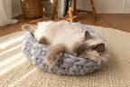 Solace Aura Pebble Grey cat bed made from natural merino wool, shown on a woven jute rug with a long-haired cat resting inside the bed. The cat is lying on its side with its head relaxed over the braided edge, paws draped forward, highlighting the soft texture and supportive shape of the hand-crafted wool bed in a calm, sunlit interior setting.